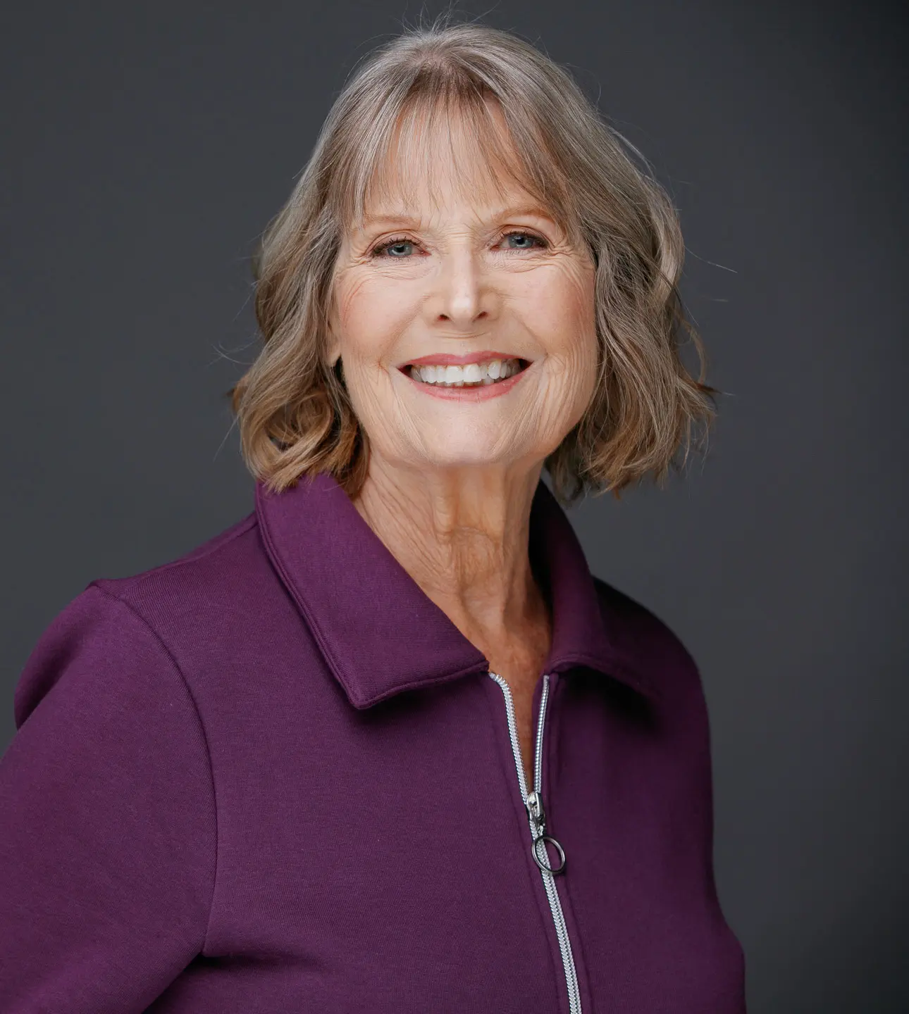 Smiling older woman in a purple jacket against a gray background.